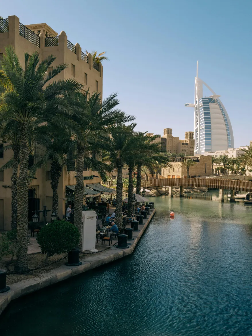 A scenic view of the sail-shaped Burj Al Arab hotel in Dubai, seen from a palm-lined canal at Madinat Jumeirah with a wooden bridge and outdoor cafe seating by the water.